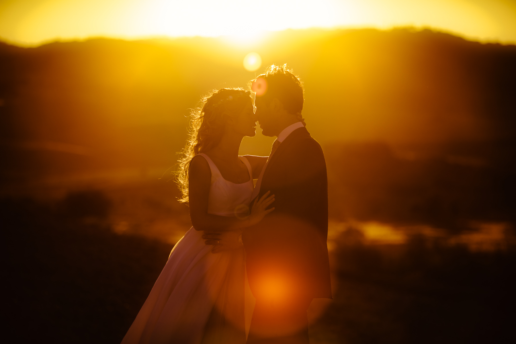 A bride and groom, Davide and Flavia, stand close together, about to kiss, silhouetted against a sunset with golden light and lens flare in the background.