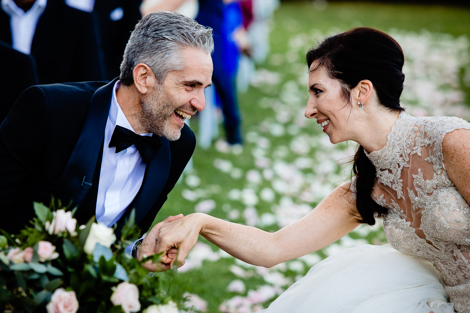 A smiling bride in a white dress holds hands with a laughing groom in a black tuxedo outdoors, surrounded by scattered flower petals on the grass—Arrigo & Ellen share a joyful moment on their special day.