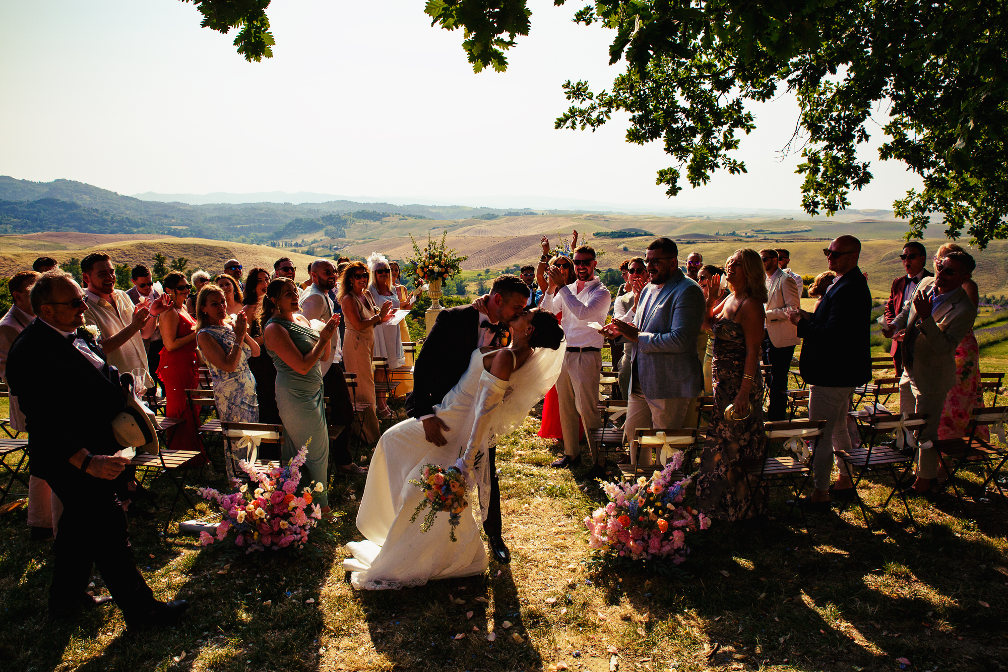 A newlywed couple shares a kiss outdoors under a tree, surrounded by clapping guests. The bride holds a bouquet and wears a white dress; the groom is in a suit. Rolling hills and bright sunlight create a scenic background.