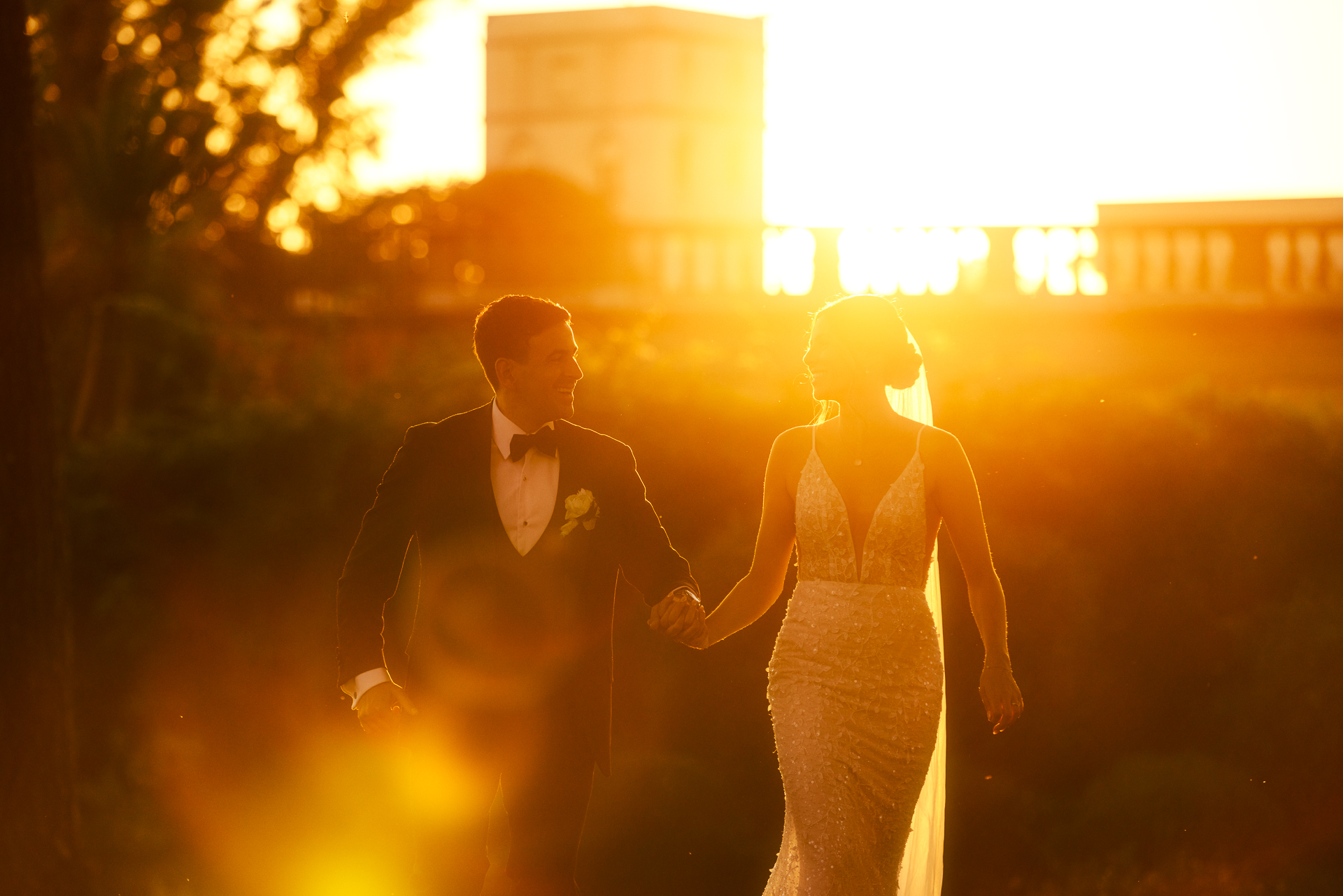 A bride and groom in formal wedding attire walk hand in hand outdoors at sunset, bathed in warm golden light, with greenery and a building blurred in the background.